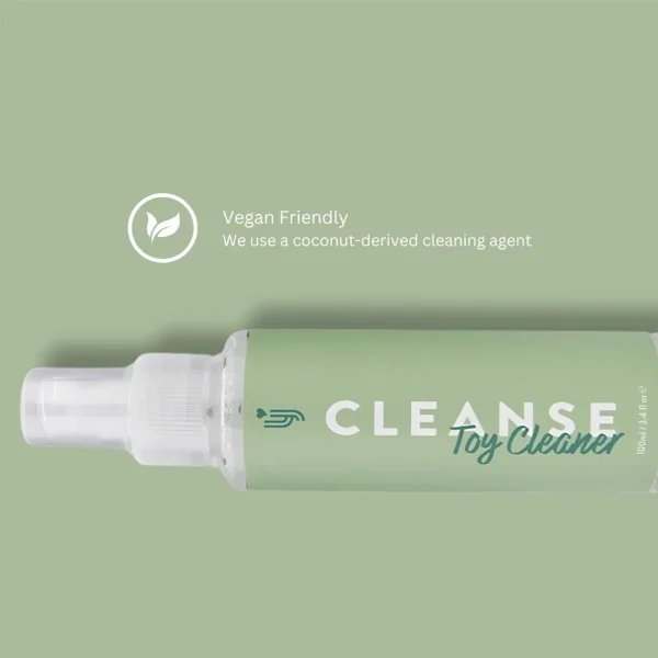 A bottle labeled "Loving Joy Cleanse Toy Cleaner" with a green label lies on a matching green background. Text reads, "Vegan Friendly. We use a coconut-derived cleaning agent," accompanied by a leaf icon above the text.