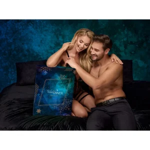 A smiling couple sits on a bed, partially dressed, opening the Feel The Magic Shiver Erotic Advent Calendar 2025 together in a dark, elegant room with a blue textured background.