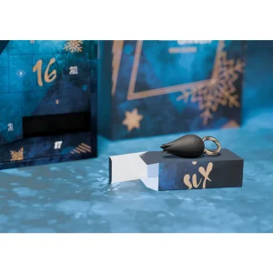 A small black teardrop-shaped item with a looped handle sits atop a blue and gold "six" box. Behind it is the Feel The Magic Shiver Erotic Advent Calendar 2025 with its door marked "16" open.