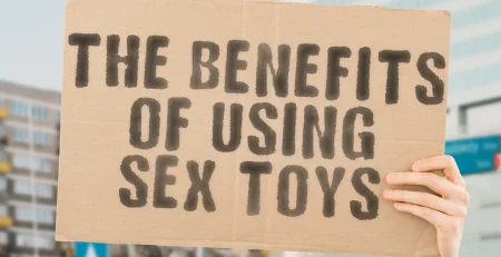 A person holds a cardboard sign with bold, black text that reads, "The Benefits of Using Sex Toys." The background is slightly blurred, showing some buildings and a street, focusing attention on the message.