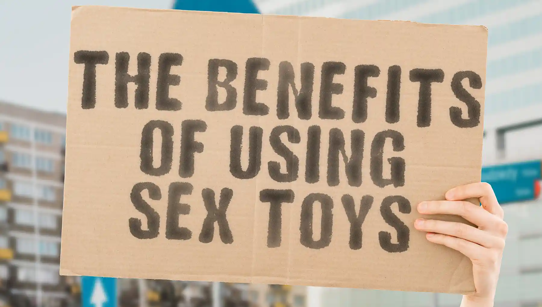 A person holds a cardboard sign with bold, black text that reads, "The Benefits of Using Sex Toys." The background is slightly blurred, showing some buildings and a street, focusing attention on the message.