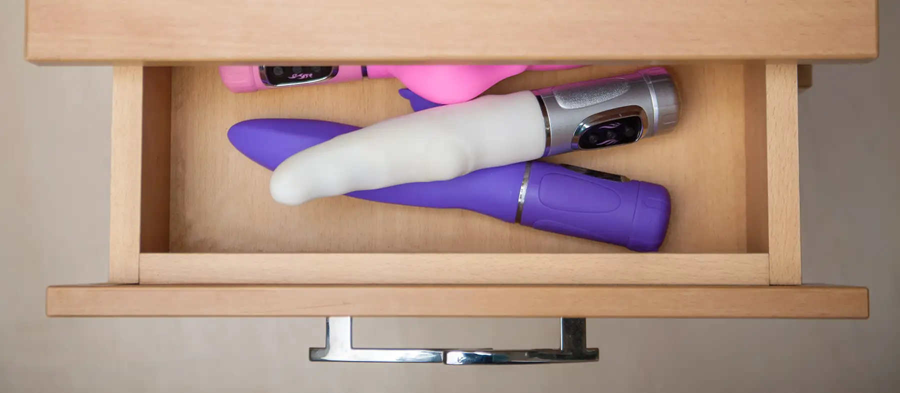 An open wooden drawer containing three sex toys of different shapes and colours, including white, purple, and pink. The handle of the drawer is metallic.