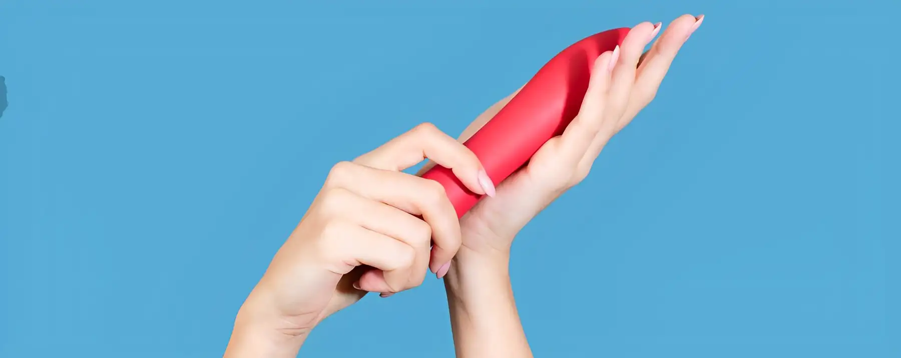 A pair of hands holding a red, sleek, elongated sex toy against a blue background. The sex toy has a smooth surface and is held gently between the fingers of one hand while the other hand supports it from below.