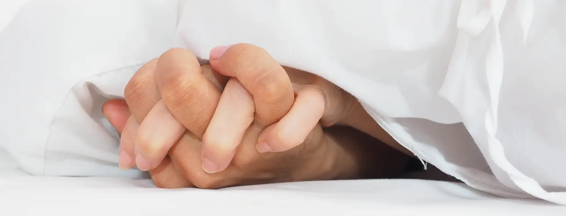 Close-up of two hands interlocked under white sheets, with fingers gently curled around each other, suggesting intimacy, connection and communication about sex toys. The white sheets partially cover the hands, creating a soft and cozy atmosphere.