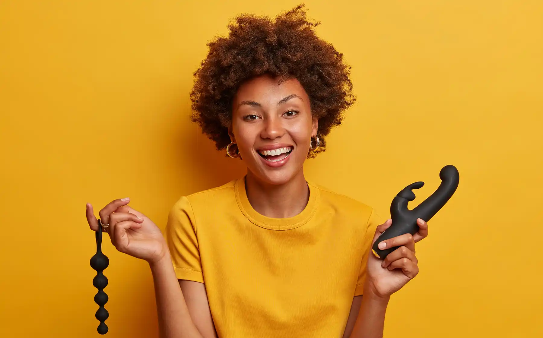 A smiling person with curly hair stands against a yellow background, holding black anal beads in one hand and a black dual-headed adult toy in the other. They are wearing a yellow T-shirt and gold hoop earrings.