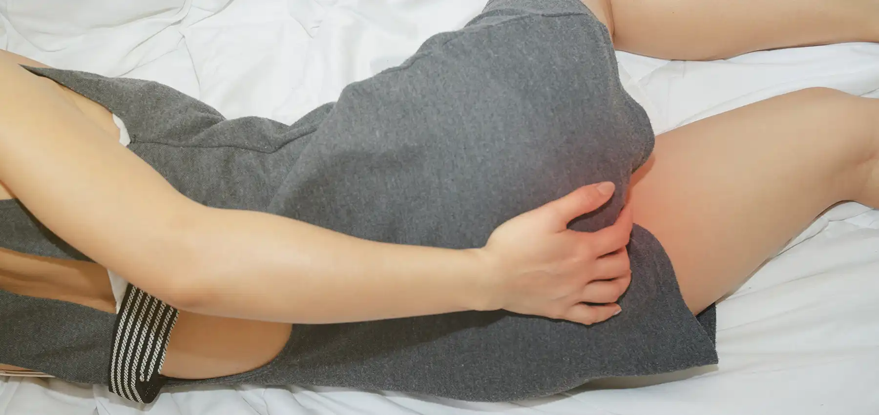A person in a grey dress lies on their side on a white bed, gently cradling their bum. The image focuses on the bum, with the hand resting on the bum highlighting pain and discomfort from anal sex.