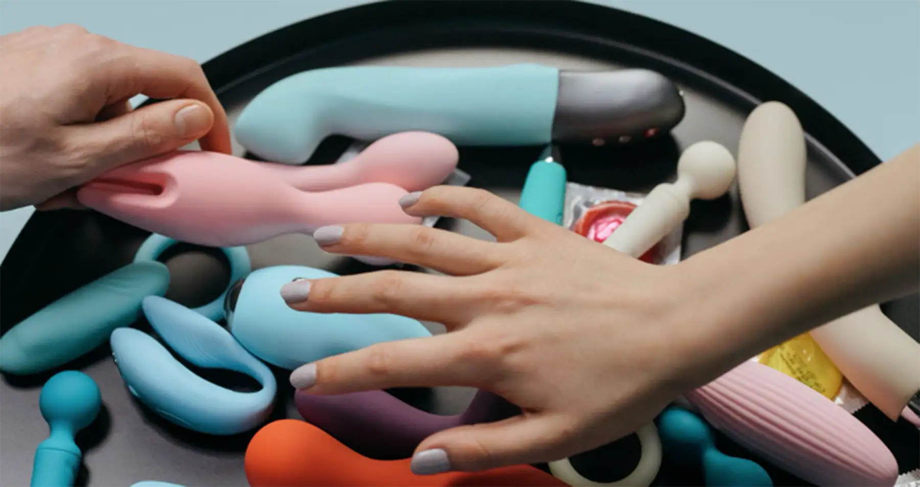 A tray containing various colorful adult toys, including vibrators and other sex toys, with two hands reaching towards them. One hand has painted nails, and each hand is interacting with a different toy on the tray.