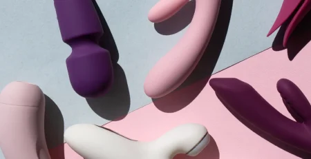A variety of sex toys in different shapes and colours are arranged on a surface divided into light blue and pink sections. The toys are predominantly shades of purple, pink, and white, with some featuring distinct curves and rounded shapes.