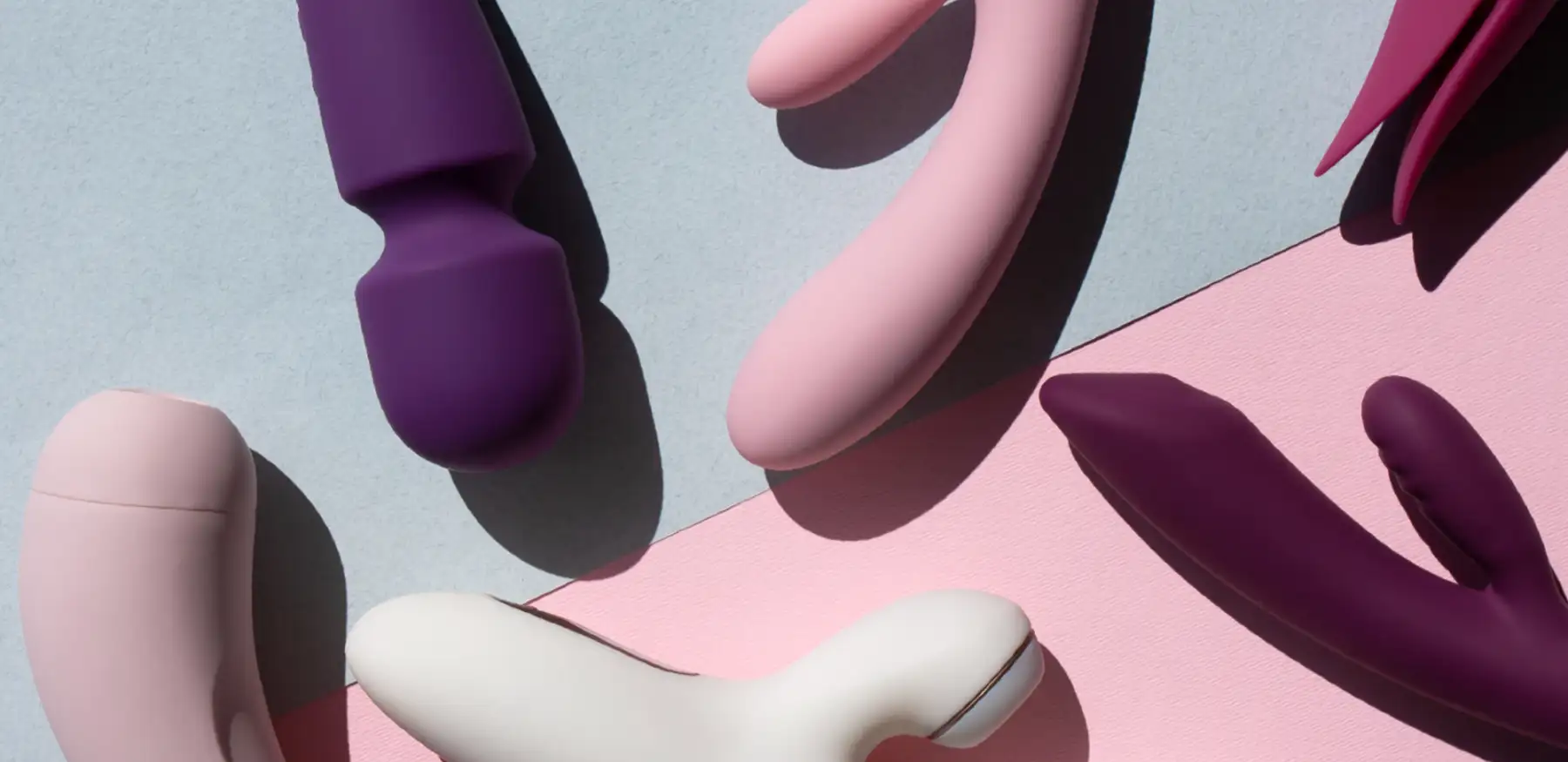 A variety of sex toys in different shapes and colours are arranged on a surface divided into light blue and pink sections. The toys are predominantly shades of purple, pink, and white, with some featuring distinct curves and rounded shapes.