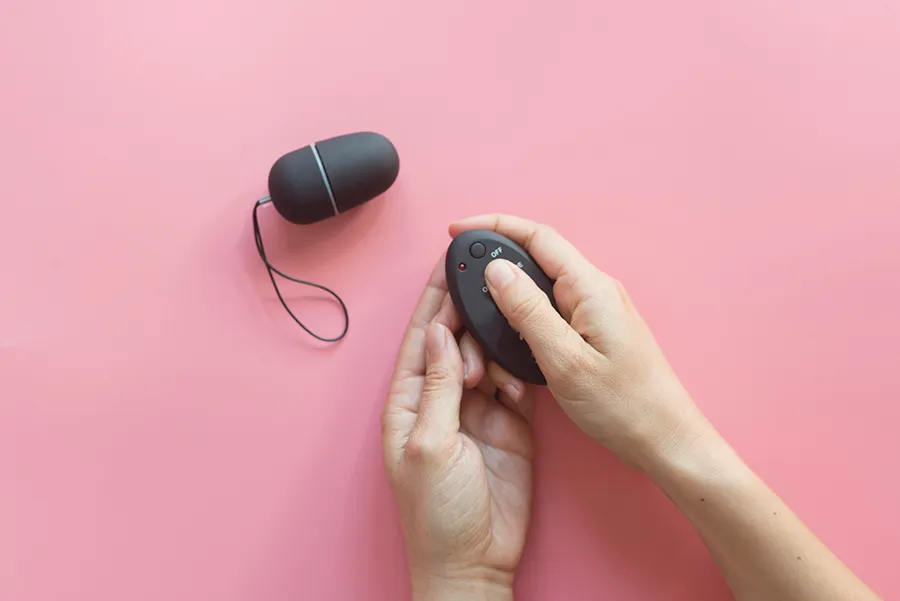 A pair of hands holding a black remote control sex toy for couples with buttons over a pink background.