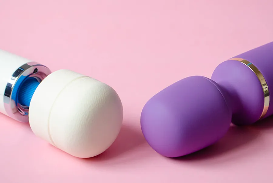 Two wand massagers are placed side by side against a pink background. The massager on the left is white with a blue accent and white rounded head, while the one on the right is purple with a silver accent and purple rounded head. Showing two sensual massage tools for couples.