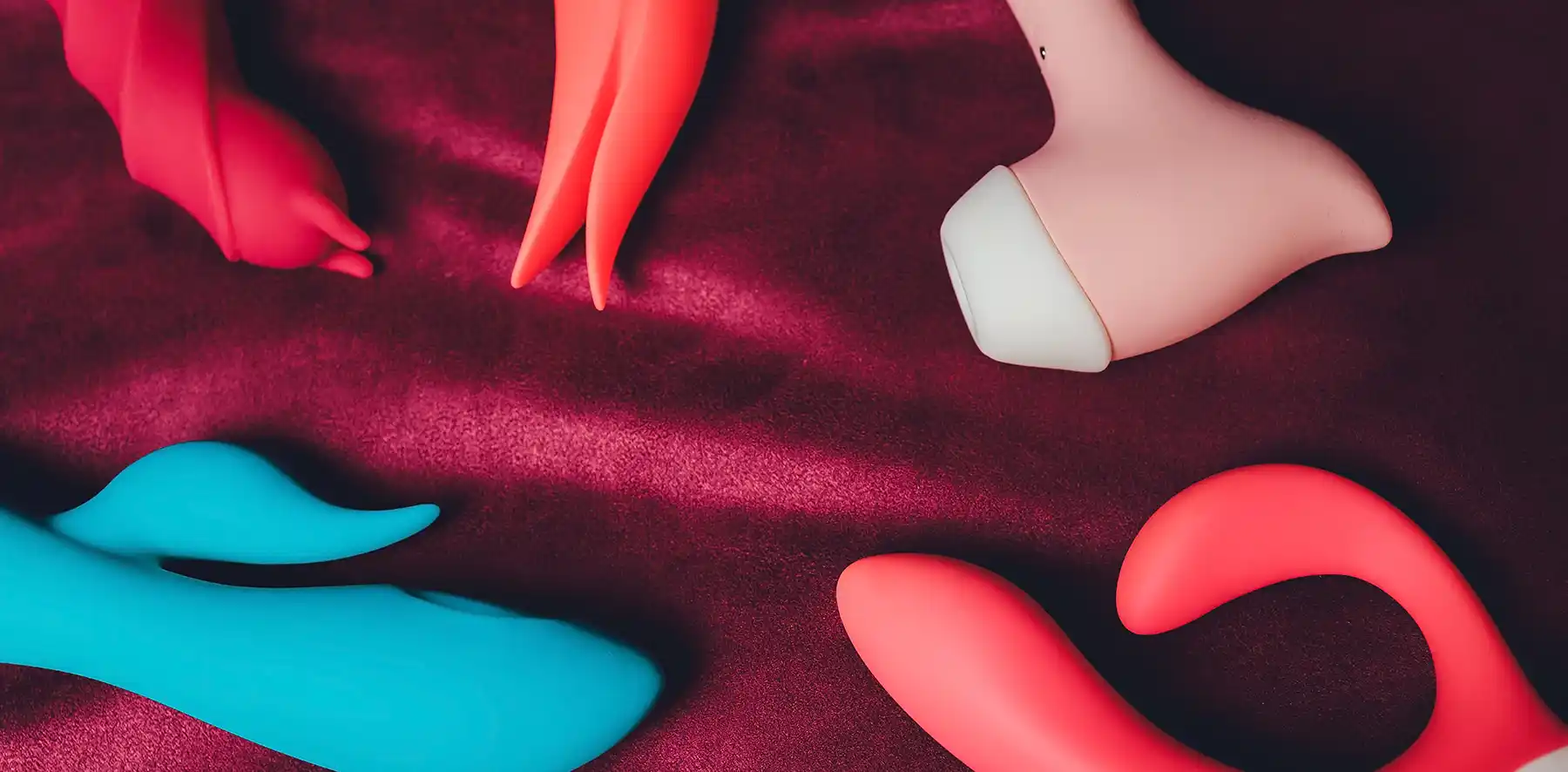 A variety of sex toys for couples in different shapes, colours, and sizes are arranged on a red fabric surface. The toys include those in blue, pink, and coral colours.