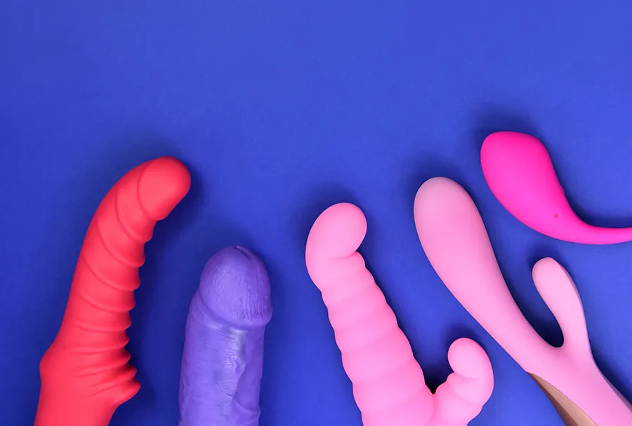 A range of colorful silicone sex toys is arranged on a blue background. The toys vary in shape, size, and color, including pink, purple, and red, with diverse designs featuring curves, ridges, and dual prongs.