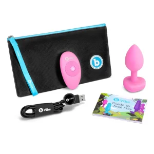 The b-Vibe Vibrating Heart Remote Controlled Butt Plug set includes a pink remote-controlled plug, black carrying case with blue zipper, USB charging cable, remote control, and a colourful "Guide to Anal Play" booklet.