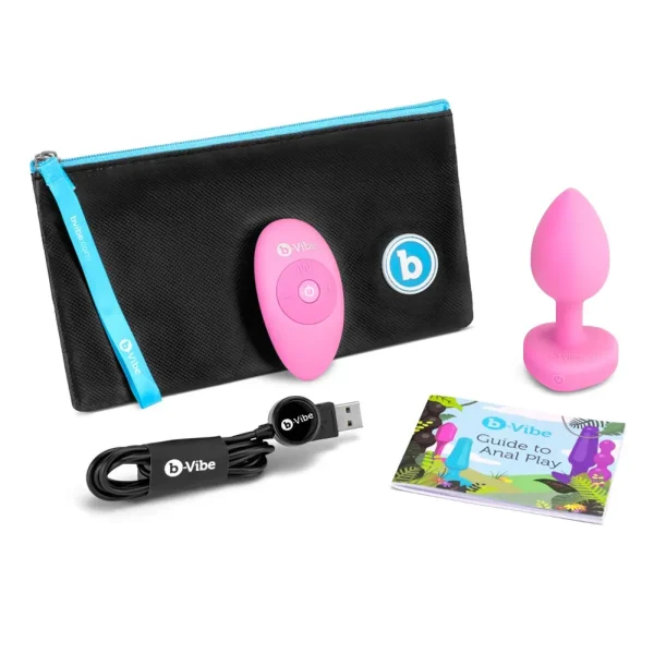 The b-Vibe Vibrating Heart Remote Controlled Butt Plug set includes a pink remote-controlled plug, black carrying case with blue zipper, USB charging cable, remote control, and a colourful "Guide to Anal Play" booklet.