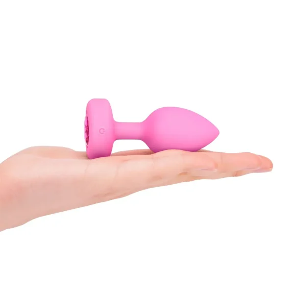 A hand holds the b-Vibe Vibrating Heart Remote Controlled Butt Plug, a small pink silicone plug with a flared base, shown against a white background.