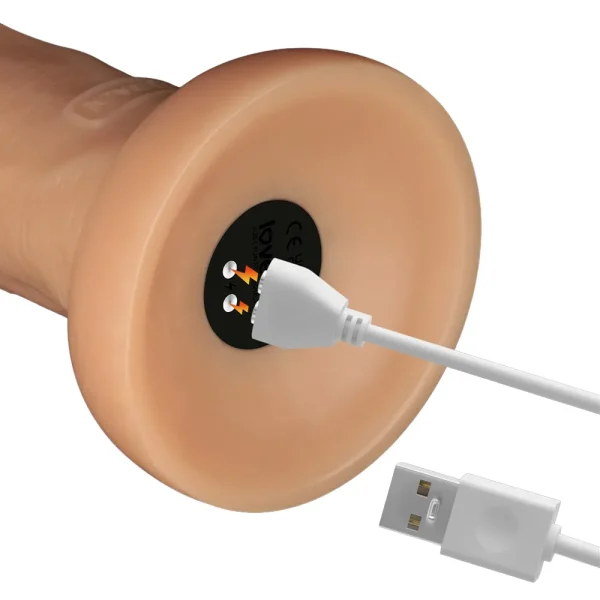 A close-up of the Power Basics 10 Inch Realistic Vibrating Dildo’s base shows it being charged with a white USB cable. The charging port is marked with lightning symbols and a CE mark.