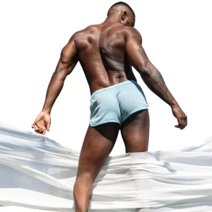 A muscular man stands outdoors with his back to the camera, wearing Sukrew Breeze Blue Net Shorts. He looks over his shoulder as sheer white fabric billows around him against a bright, white background.