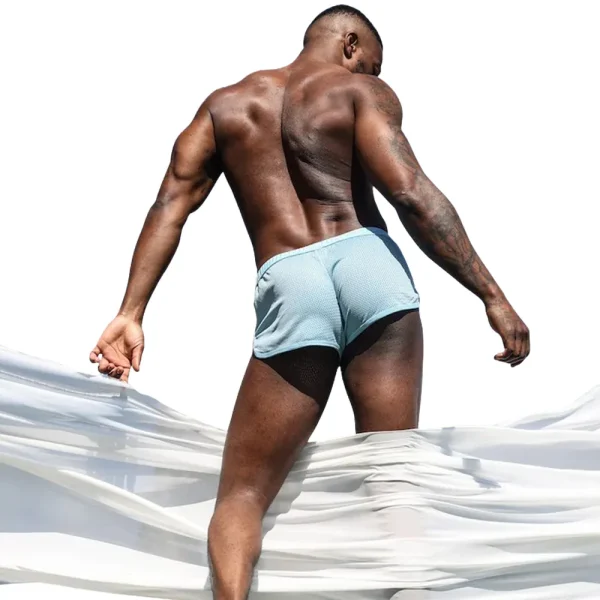 A muscular man stands outdoors with his back to the camera, wearing Sukrew Breeze Blue Net Shorts. He looks over his shoulder as sheer white fabric billows around him against a bright, white background.