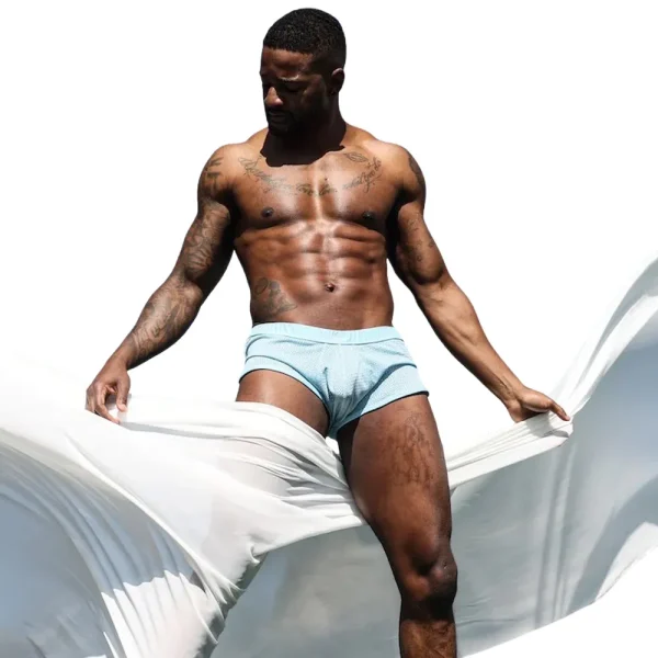 A muscular man models Sukrew Breeze Blue Net Shorts against a white background, stretching a piece of white fabric wrapped around his lower body.