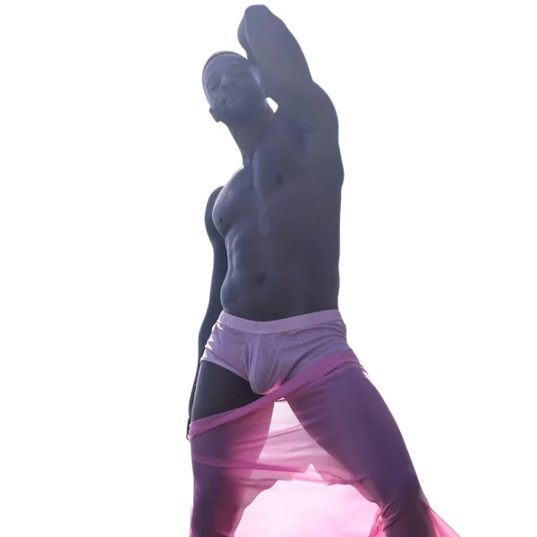 A shirtless man stands confidently against a white background, wearing Sukrew Summer Pink Net Shorts and draped with a sheer pink fabric, with one arm raised and the other by his side.