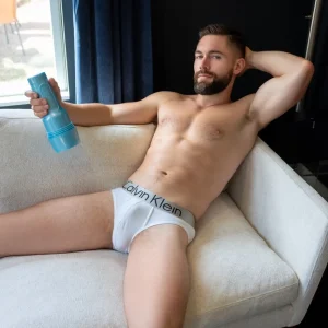 A bearded Griffin Barrows in white Calvin Klein underwear lounges on a white couch, leaning back with one arm behind his head and holding the Fleshjack Boys Griffin Barrows Cake Butt Masturbator in his other hand.