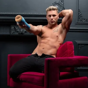 A shirtless, tattooed Josh Moore reclines on a red velvet armchair before a dark ornate wall, flexing an arm behind his head while holding the Fleshjack Boys Josh Moore Cheeky Butt Masturbator toward the camera.