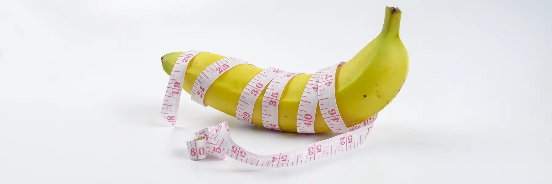 A yellow banana wrapped with a pink and white measuring tape, set against a plain white background, implying using a measuring tape to measure for a cock ring.