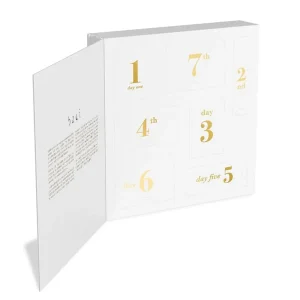 An open Baci 7 Day Hosiery Adventure Gift Box displays its white compartments numbered day one to seven in gold, arranged in a grid. The inside lid features printed text and the "Baci" brand name. Behind each door is some baci lingerie or accessories.