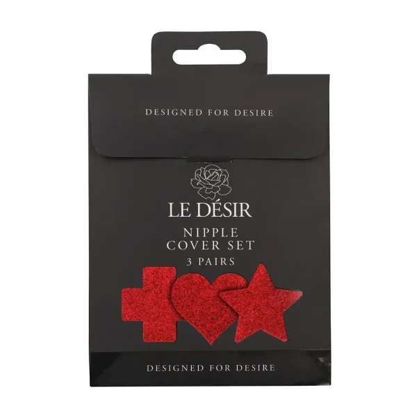 The Le Desir Nipple Cover Set comes in black packaging with "LE DÉSIR Nipple Cover Set, 3 pairs" and displaying three red glitter shapes—cross, heart, and star. The box features "DESIGNED FOR DESIRE" at the top and bottom.