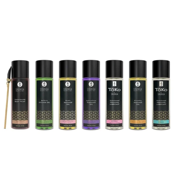 Seven bottles from Shunga Calendar of Desires. It features oils and lubricants stood side by side on a white background, with a small applicator brush next to the leftmost bottle. Each bottle features a distinctively coloured label.