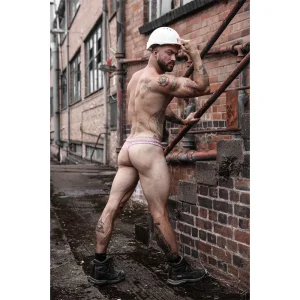 A muscular man wearing a white hard hat, black boots, and the Sukrew Diamond Classic Thong poses outdoors beside a brick building and metal pipes, flexing his arm and glancing over his shoulder.