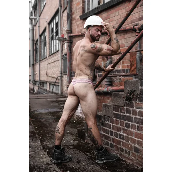 A muscular man wearing a white hard hat, black boots, and the Sukrew Diamond Classic Thong poses outdoors beside a brick building and metal pipes, flexing his arm and glancing over his shoulder.