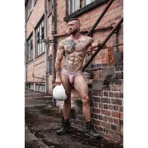 A muscular, tattooed man in a Sukrew Diamond Classic Thong, black boots, and holding a white hard hat stands outdoors by a brick building with pipes, looking into the distance.