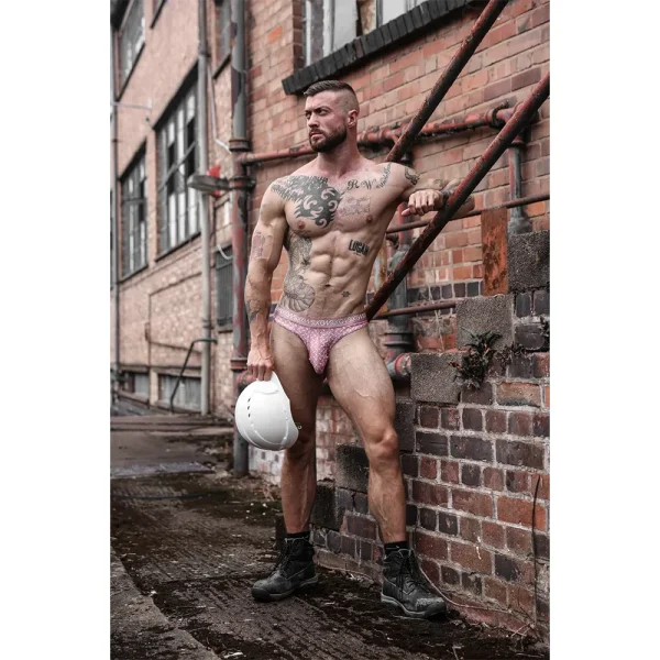 A muscular, tattooed man in a Sukrew Diamond Classic Thong, black boots, and holding a white hard hat stands outdoors by a brick building with pipes, looking into the distance.