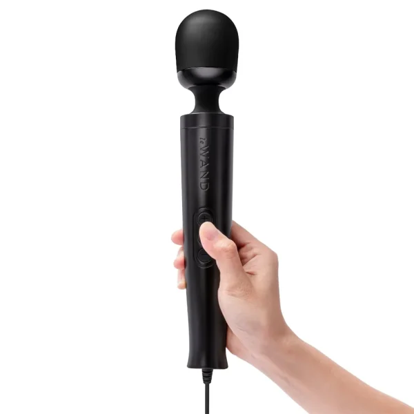 A hand holds the Le Wand Diecast Plug In Vibrating Wand Massager, featuring a black finish, rounded head, and control buttons on the handle, set against a plain white background.