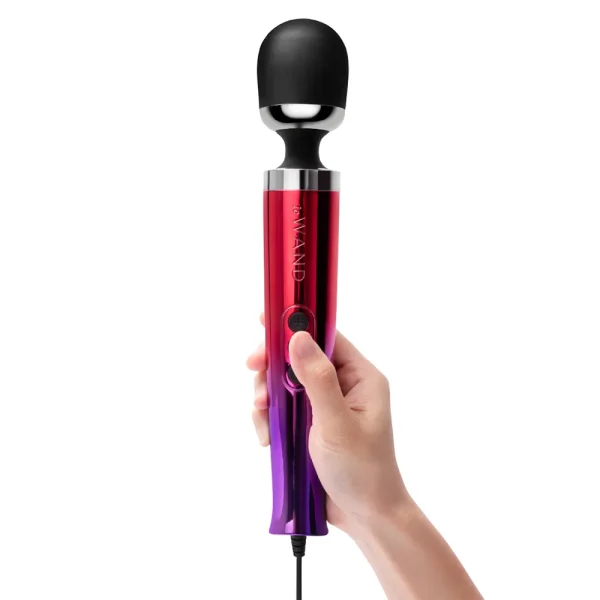 A hand holds the Le Wand Diecast Plug In Vibrating Wand Massager in ombre colour, a mains-powered device featuring a black rounded head with red and purple accents.
