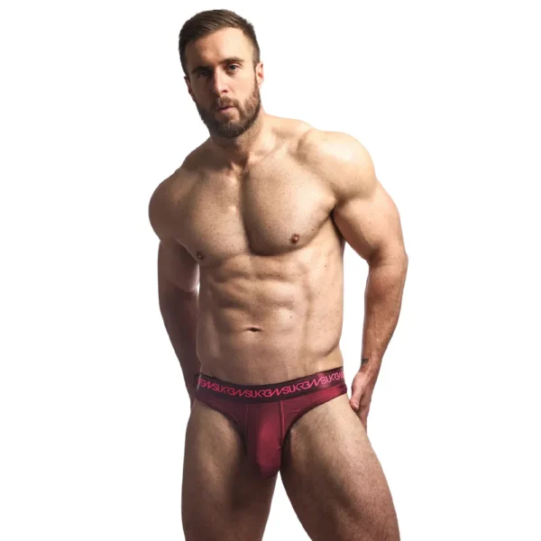 A muscular bearded man poses bare-chested, wearing only the Sukrew Clavet Classic Thong in claret red, standing against a plain white background.