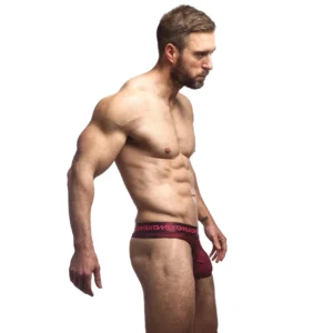 A muscular, bearded man with short hair stands in profile on a plain white background, wearing the maroon Sukrew Clavet Classic Thong.