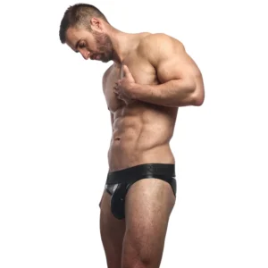 A muscular man with short hair stands in profile against a plain white background, wearing the Sukrew Fetish Black Wet Look Classic Brief, one hand on his chest and looking down.