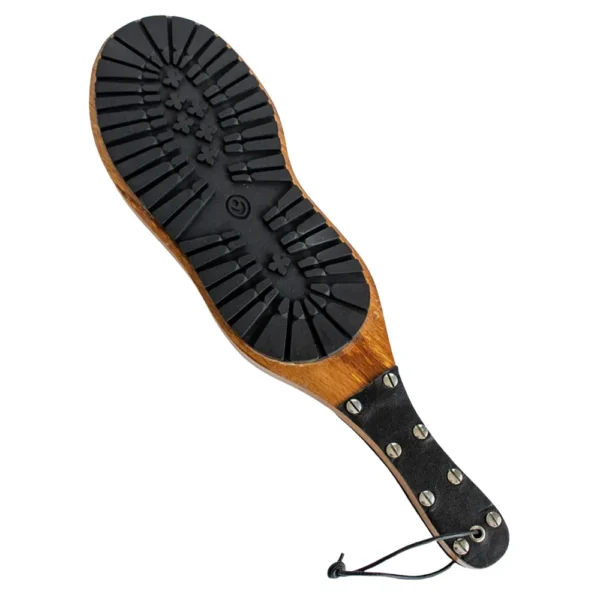 wooden-boot-imprint-paddle The Wooden Boot Imprint Paddle features a black faux leather-wrapped handle with metal studs and a rubber sole with deep treads on its flat wooden surface.