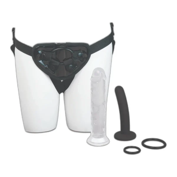 me-you-us-complete-harness-kit-detail-3 A strap-on harness is displayed on a mannequin’s hips, alongside a transparent dildo, a black dildo, and two O-rings, all against a white background.