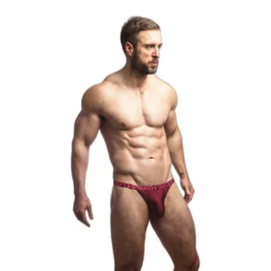 A muscular man with a beard stands against a white background, wearing only a red thong with a patterned waistband. He looks off to the side with a serious expression.