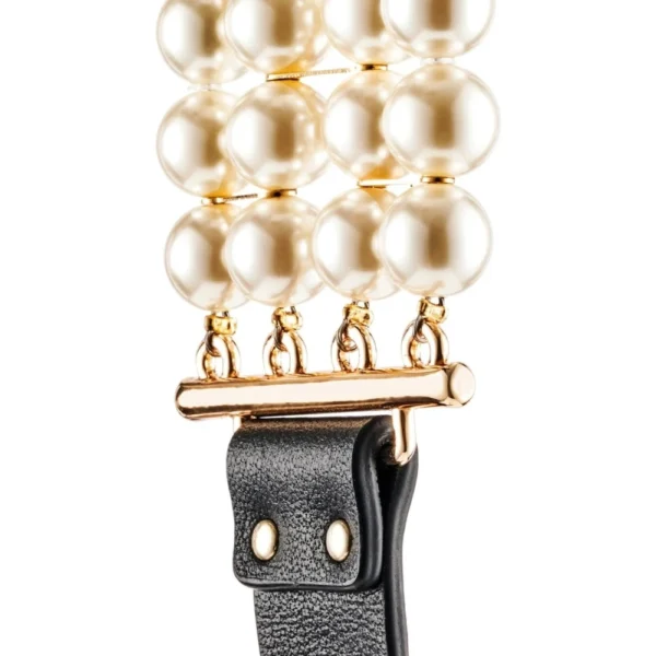 Close-up of a watch strap featuring three rows of large, glossy white pearls connected by a gold bar, attached to a textured black leather band with silver studs.