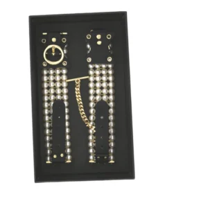 A black tray holds a pair of decorative black leather cuffs adorned with silver beads and connected by a gold chain and ring. The cuffs are arranged neatly side by side.