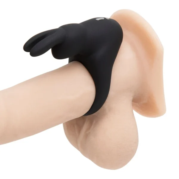 A black silicone ring shaped like a bunny is placed on a realistic model of a penis and testicles, shown in close-up against a white background.