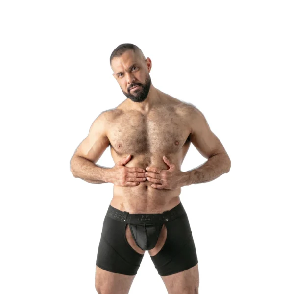 A muscular man with a beard poses bare-chested, wearing black boxer shorts with a large front cut-out, standing against a plain white background.