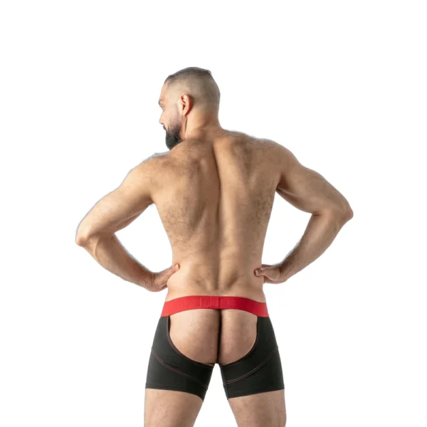 A muscular, bearded man with a shaven head stands with hands on hips, facing away, wearing black boxer shorts with a red waistband and an open back, exposing his buttocks. The background is plain white.