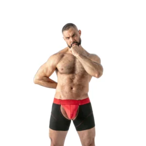 A muscular, bearded man stands on a white background wearing black and red boxer shorts with a front cut-out, resting one hand on his hip and the other under his chin in a thoughtful pose.