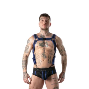 A tattooed person stands facing forwards, wearing a black and blue harness and matching underwear, against a plain white background.